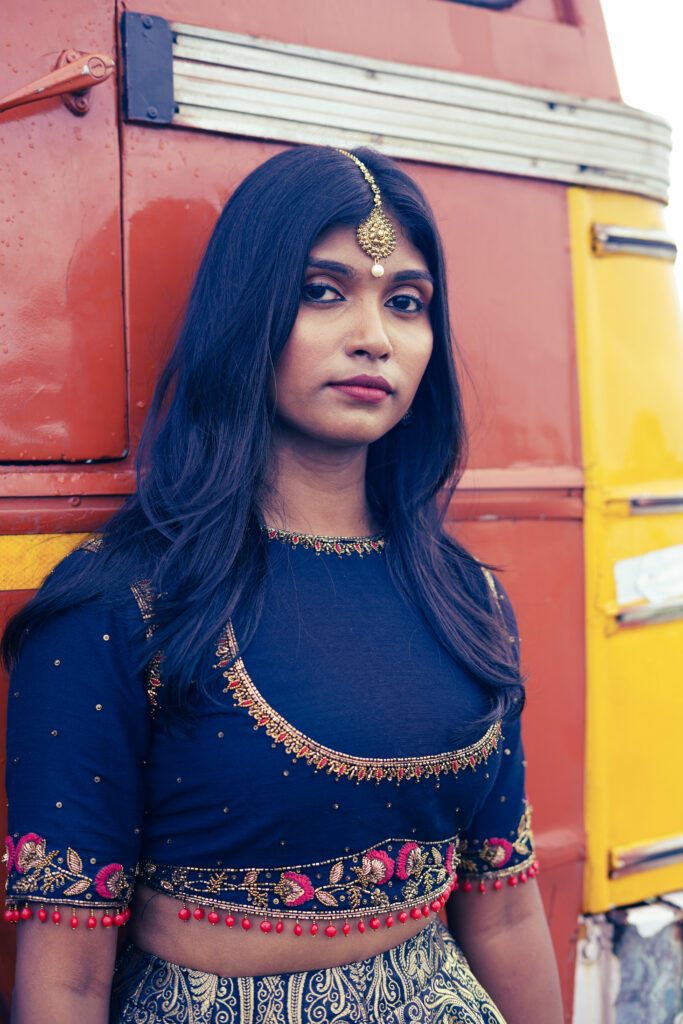 Portrait of a woman wearing a maang tikka and navy embroidered blouse against a red and yellow bus