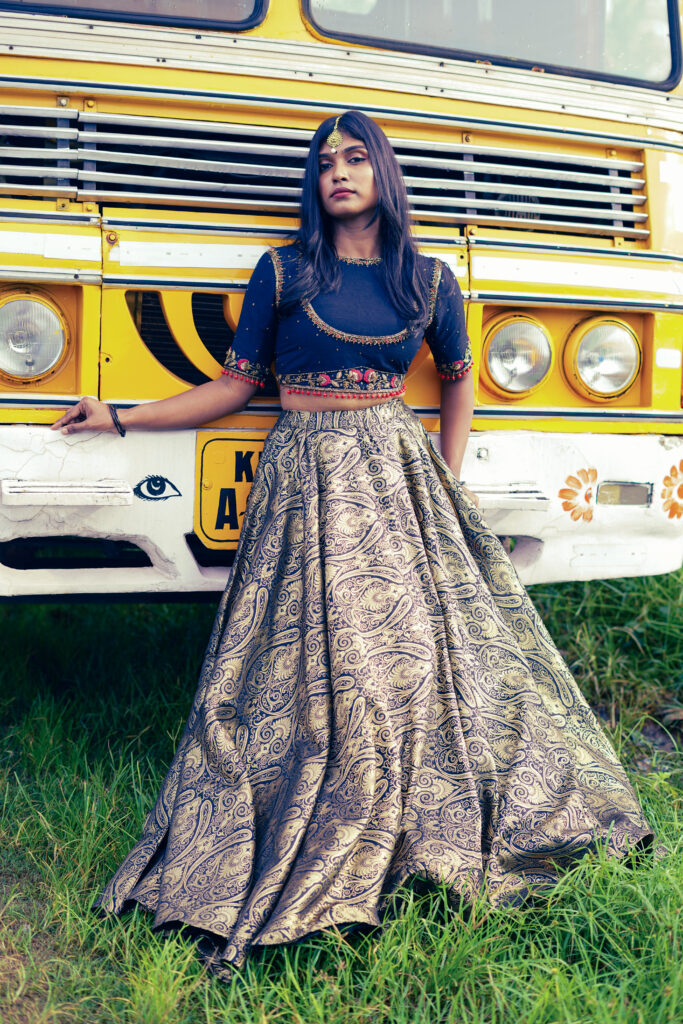 Woman in a navy embroidered blouse and gold lehenga posing in front of a yellow Kerala bus