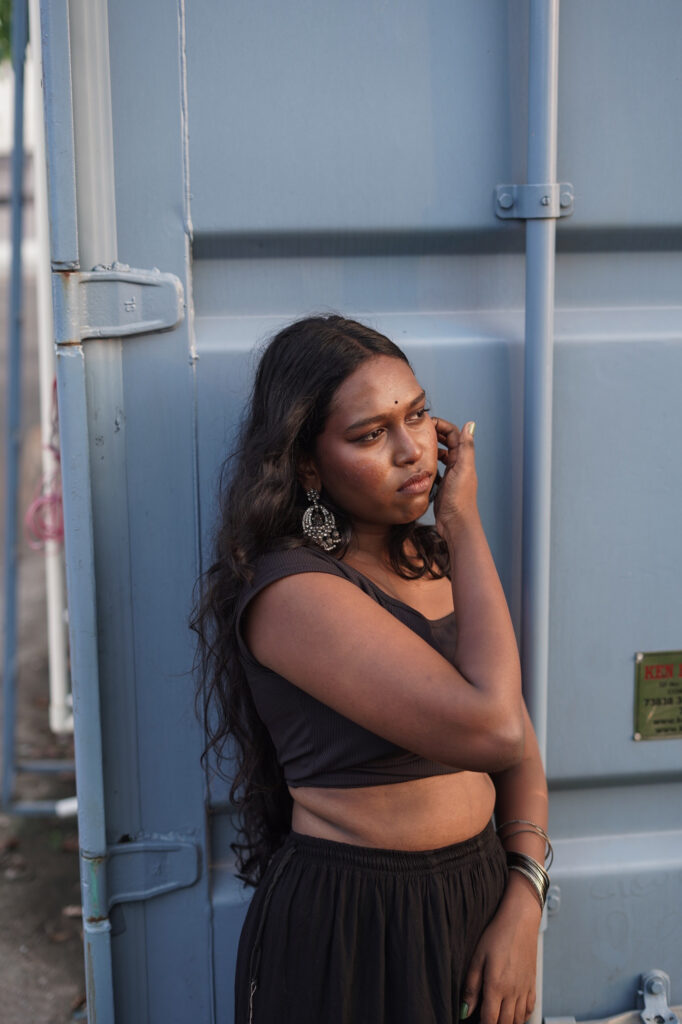 Woman in a black outfit and jhumka earrings against a blue shipping container