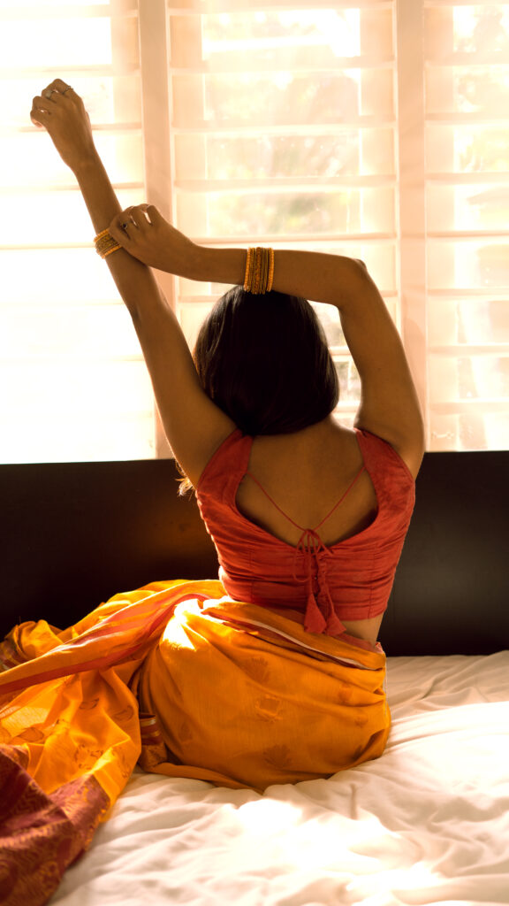 Woman in an orange saree stretching by a bright window, seen from behind