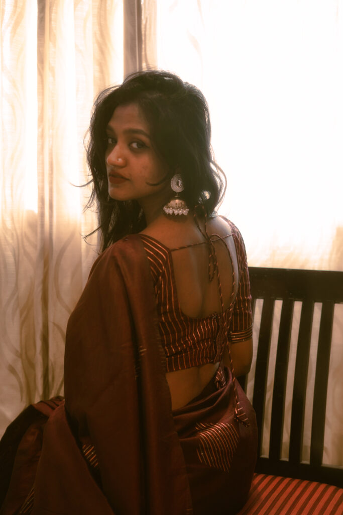Woman in a brown saree seated by a bright window, looking back