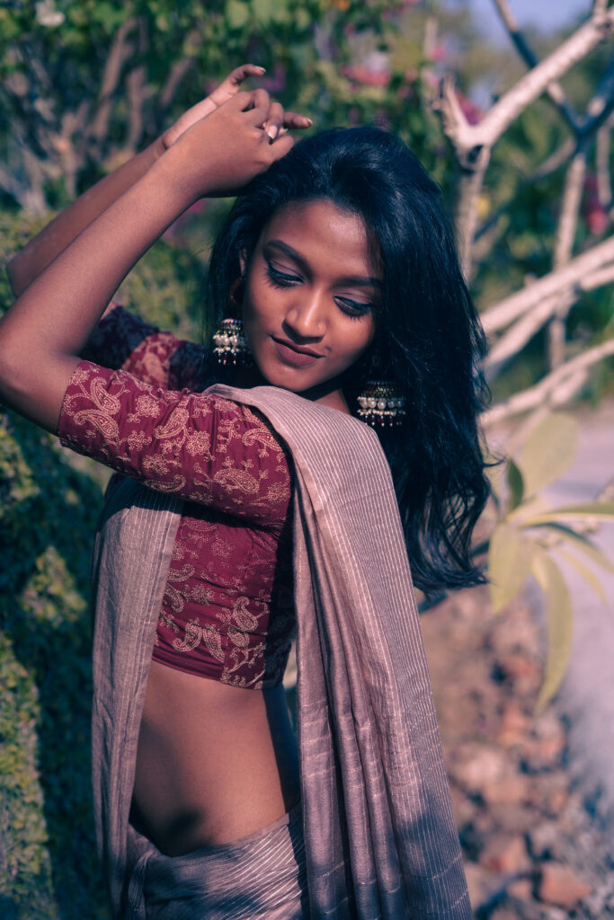 Woman with her arms raised, wearing a red printed blouse and grey saree in warm light