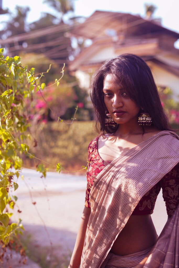 Woman in a striped saree and jhumkas beside a traditional tiled-roof Kerala house