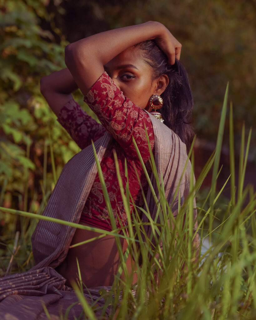 Woman posing in tall grass wearing jhumka earrings and a red printed blouse, warm light