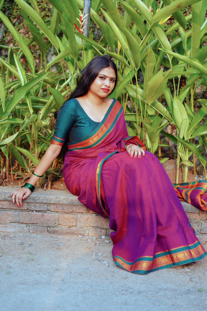 Woman in a magenta and green silk saree seated in front of tall leaves