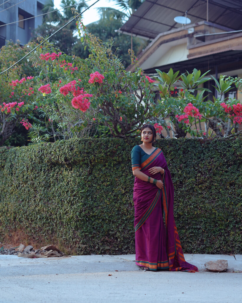 Wide portrait of a woman in a magenta saree framed by red flowers and a garden hedge