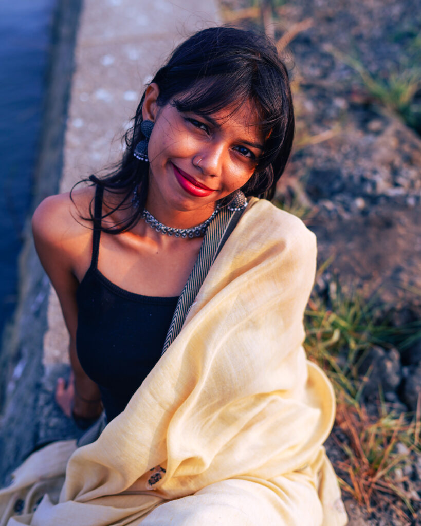 Smiling woman in a cream saree sitting on riverside rocks