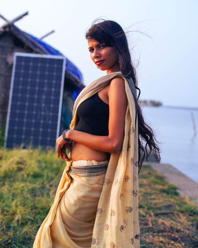 Woman in a cream saree and black blouse standing by a river bank, hair lifted by the wind