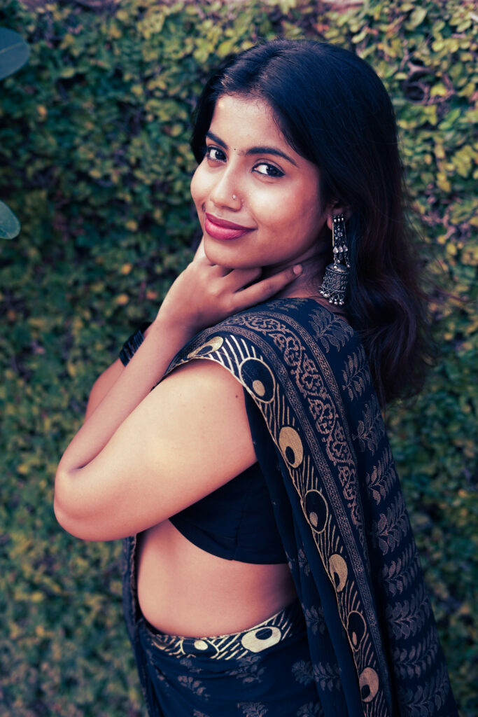 Smiling woman in a black and gold saree against a green wall