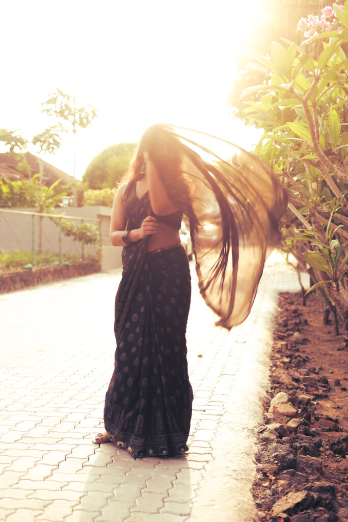 Woman swirling her saree pallu on a sunlit path lined with flowers