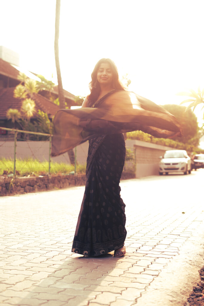 Woman in a black printed saree on a sunlit paved path, saree catching the light