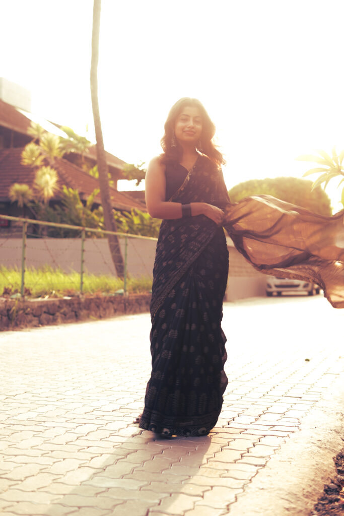 Woman in a black printed saree on a sunlit paved path, saree catching the light