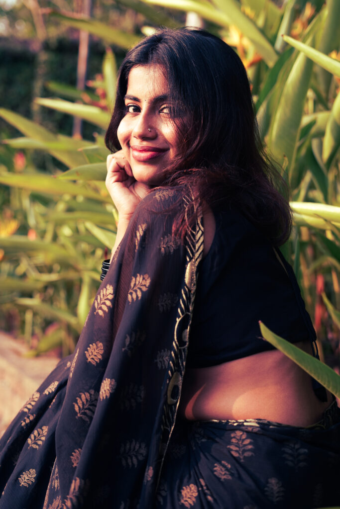 Smiling woman in a black and gold saree against a green wall