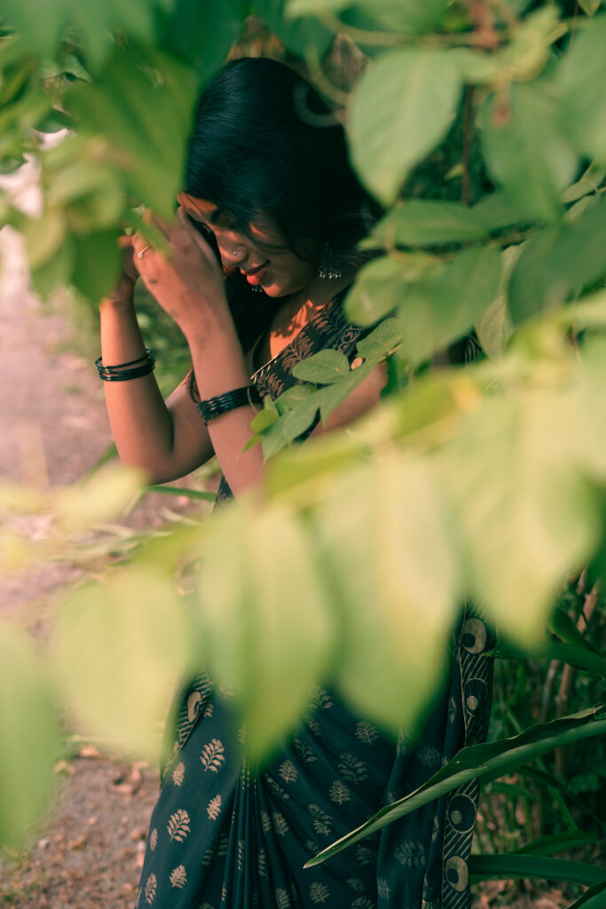 Woman in a printed saree partly hidden by green leaves in the foreground