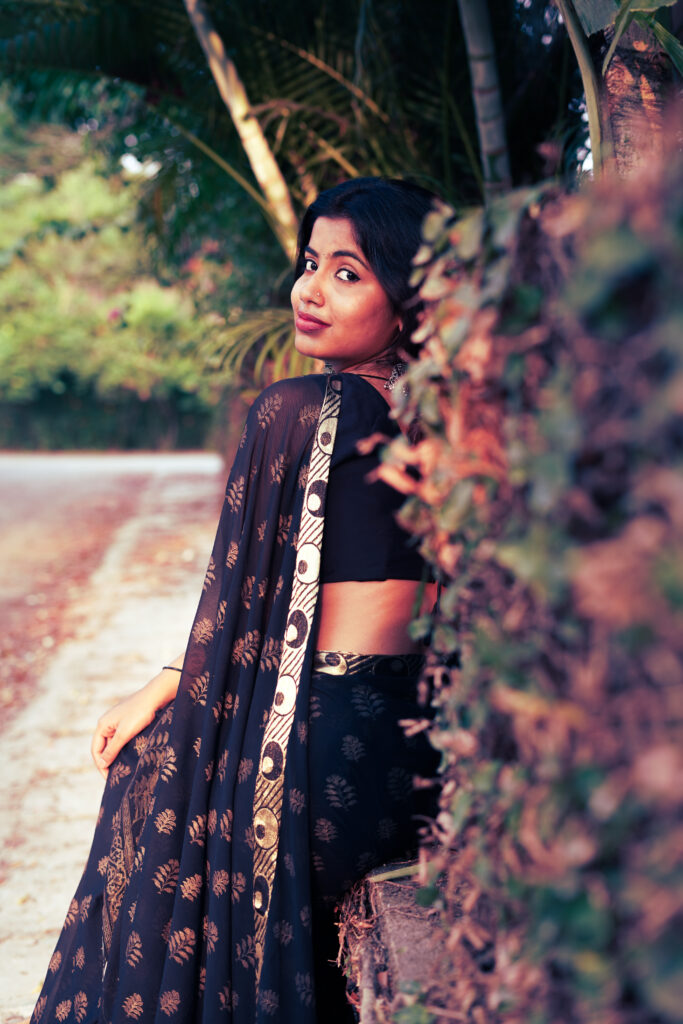 Woman in a black printed saree looking back beside a green hedge