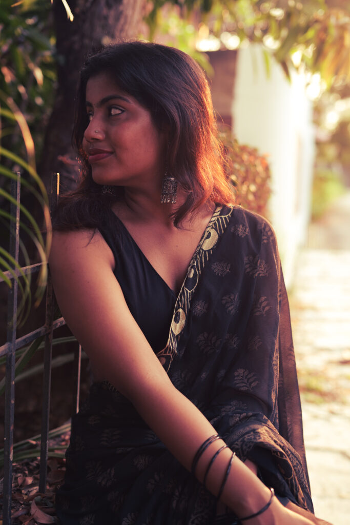 Woman in a black printed saree seated in warm evening light among leaves