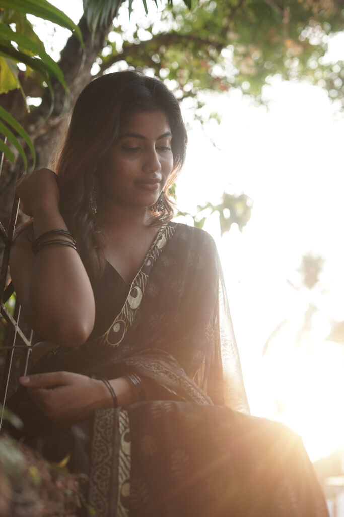 Woman in a black printed saree seated in warm evening light among leaves