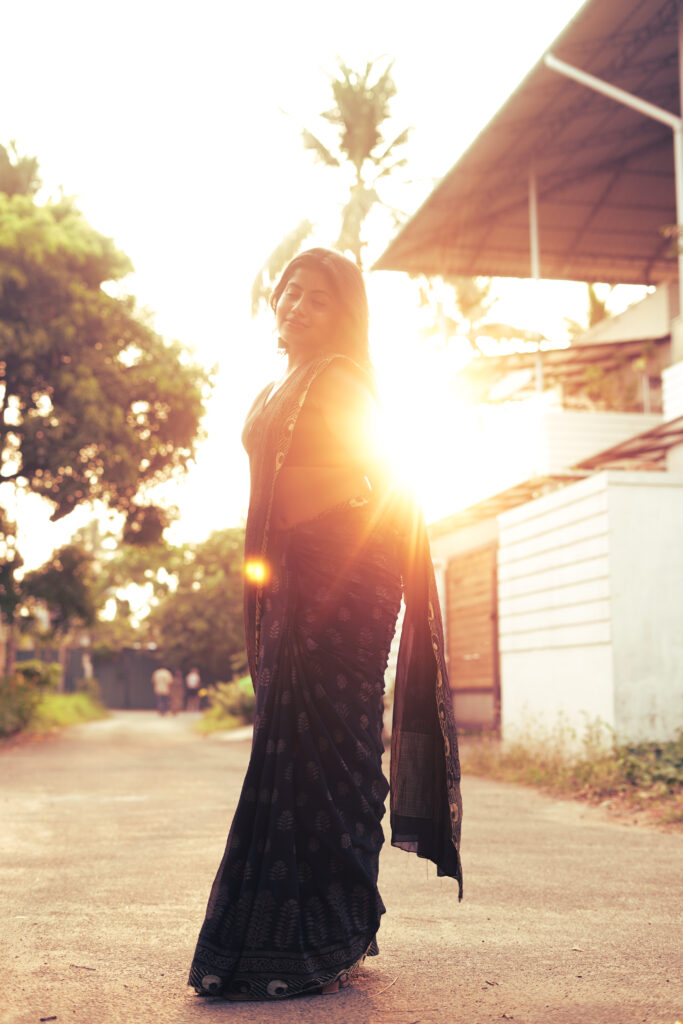 Woman in a black printed saree walking into golden-hour sunlight on a quiet village road