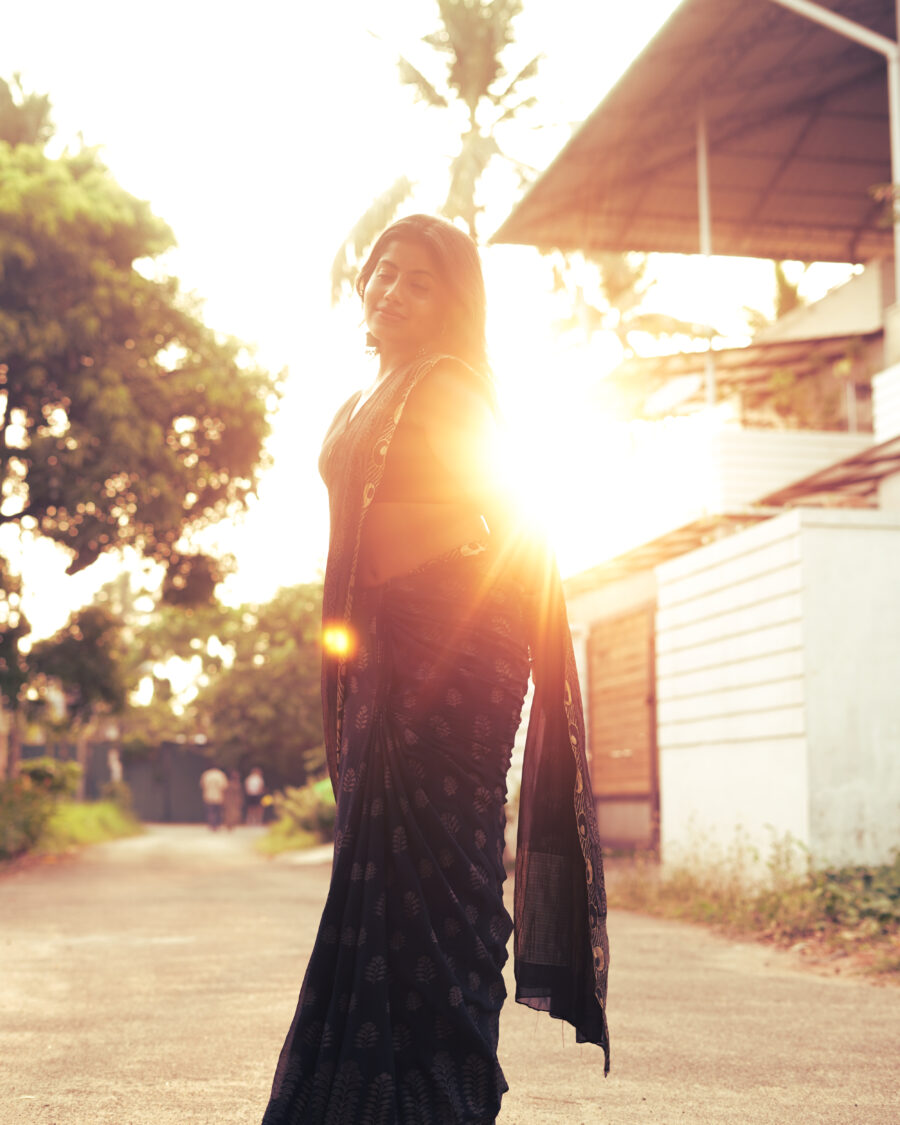 Woman in a black printed saree walking into golden-hour sunlight on a quiet village road