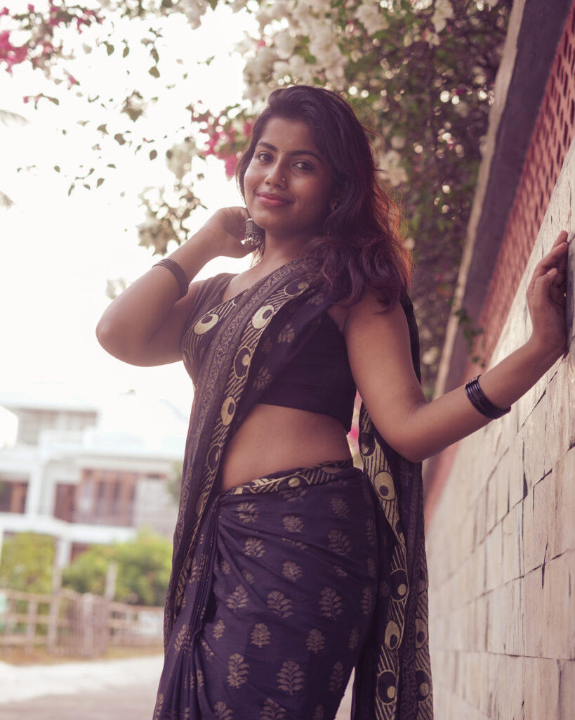 Woman in a black printed saree leaning against a wall under pink bougainvillea
