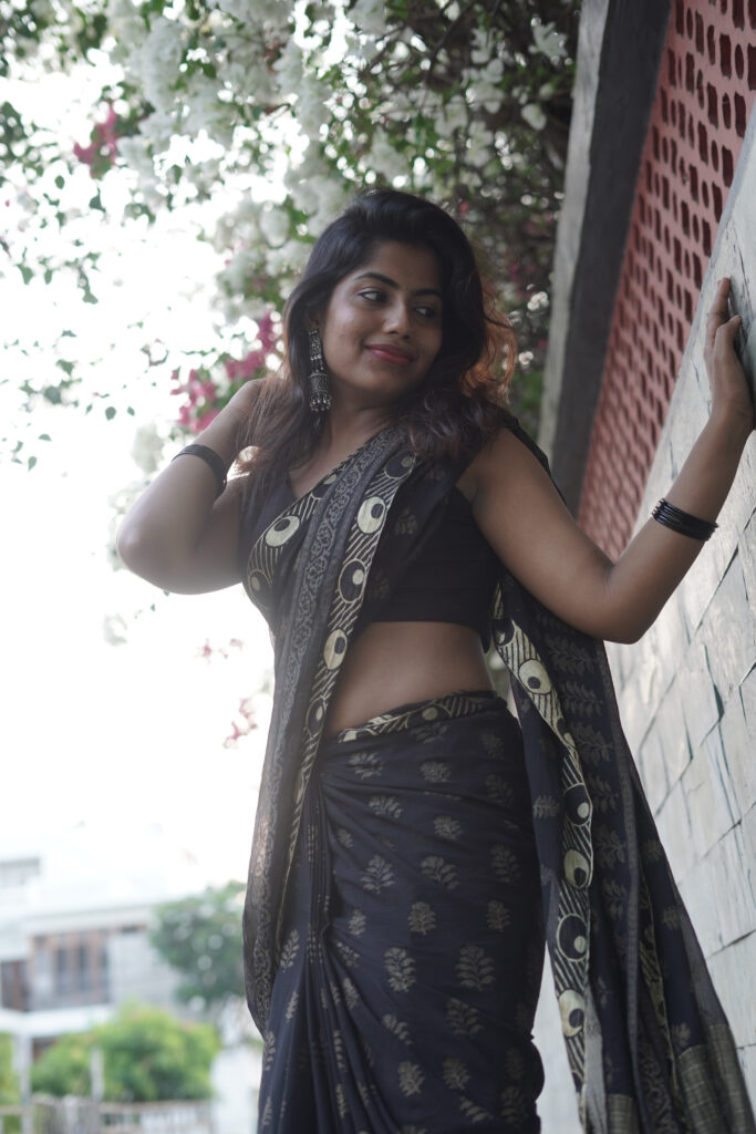Woman in a black printed saree leaning against a wall under pink bougainvillea