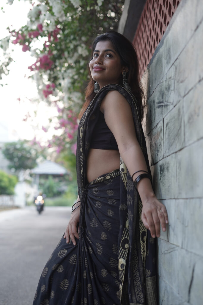 Woman in a black printed saree leaning against a wall under pink bougainvillea