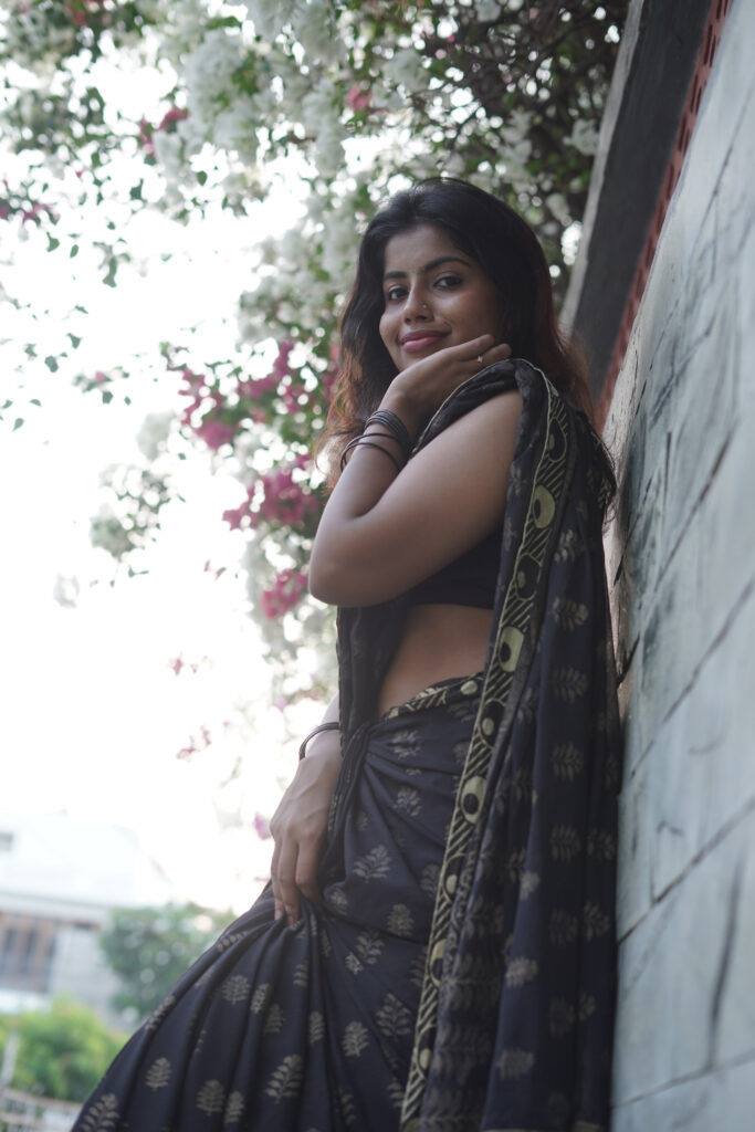 Woman in a black printed saree leaning against a wall under pink bougainvillea