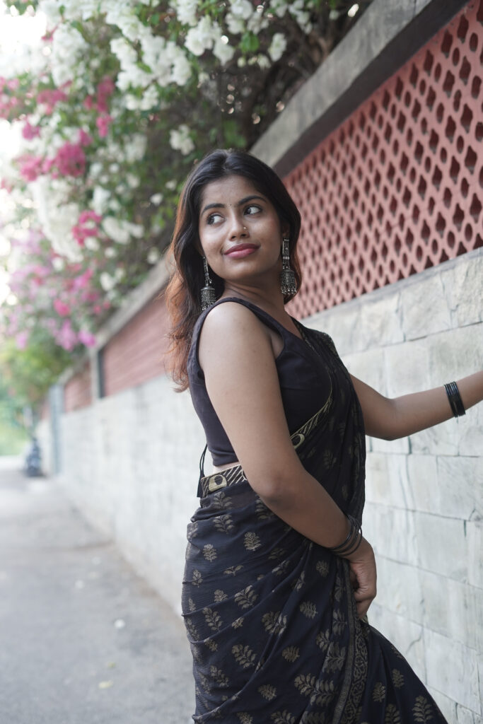 Woman in a black printed saree leaning against a wall under pink bougainvillea