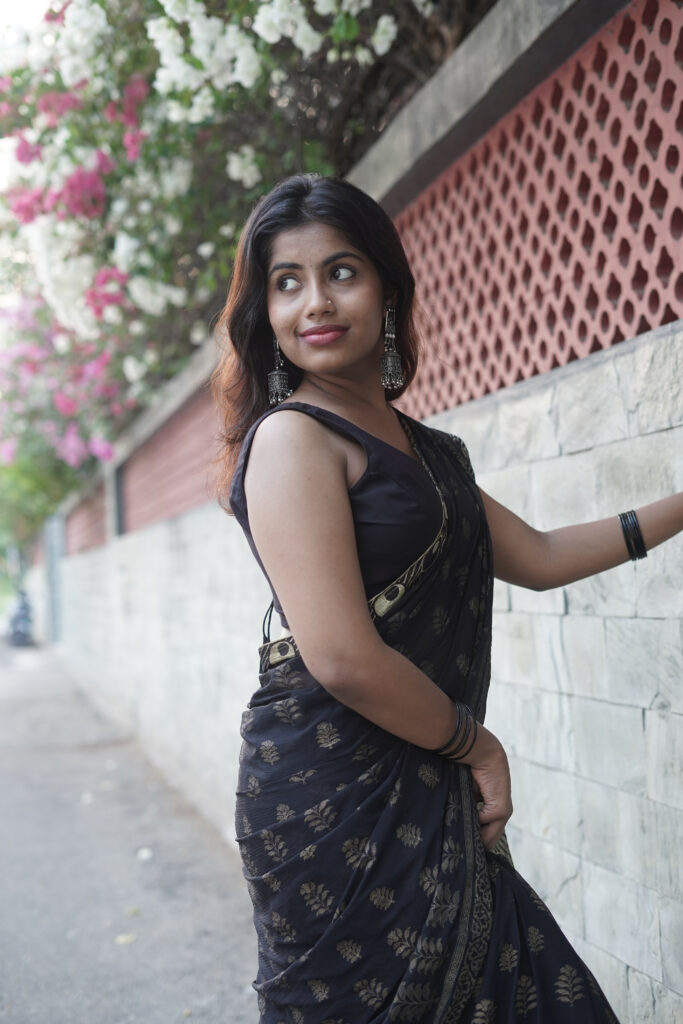 Woman in a black printed saree leaning against a wall under pink bougainvillea