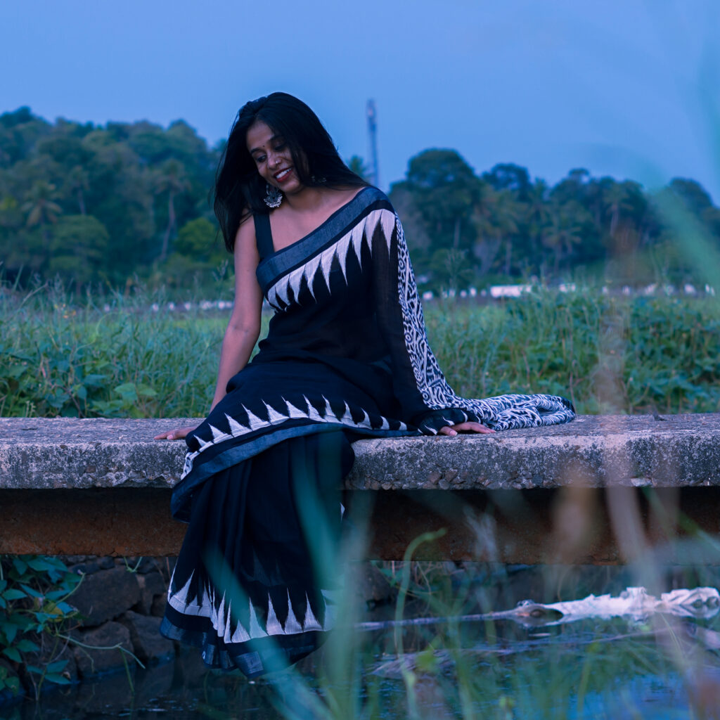 Woman in a black and white saree seated on a low wall in a green field at blue hour