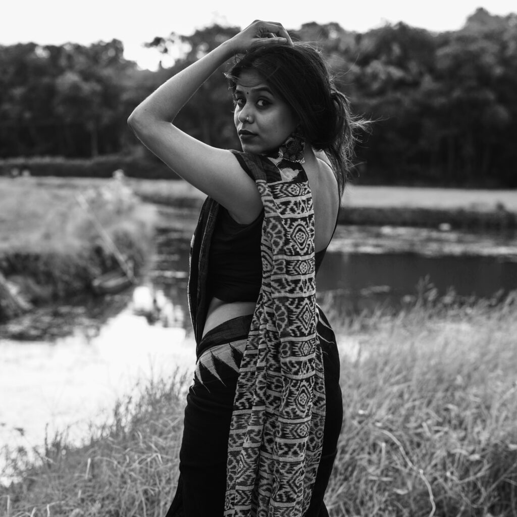 Black and white portrait of a woman in a printed saree beside water and tall grass