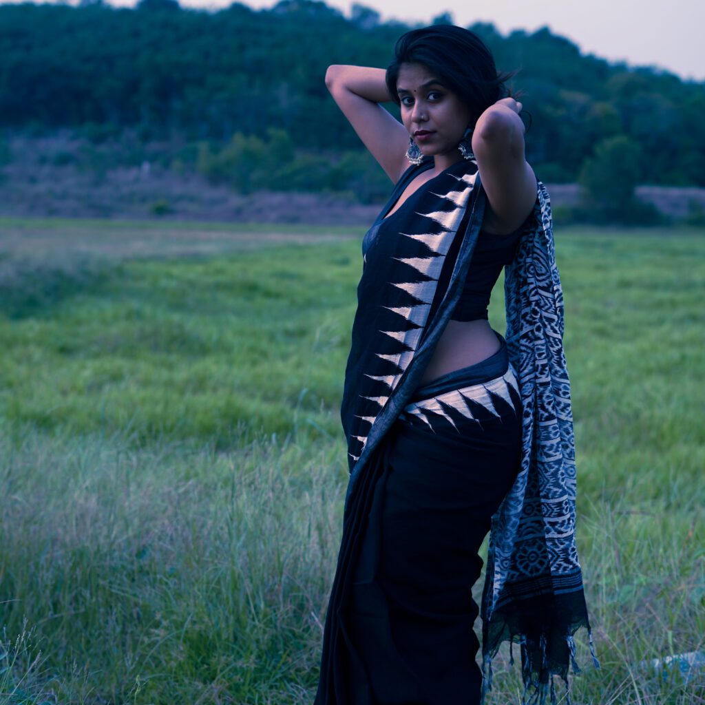 Woman in a black and white printed saree standing in a grassy field at dusk