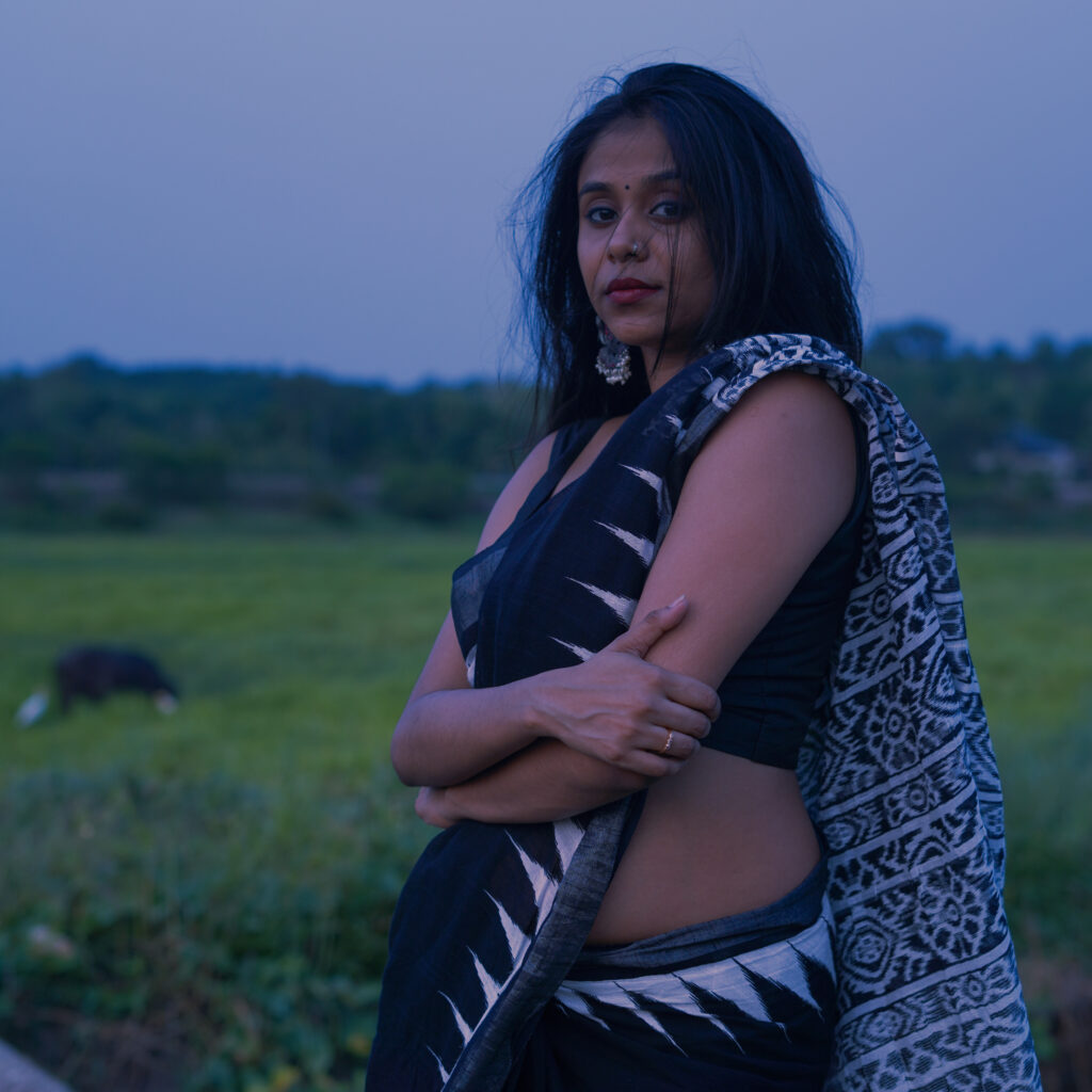 Woman in a black and white saree with arms crossed, green fields behind her at dusk