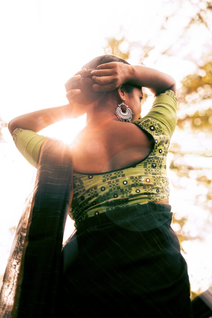 Backlit portrait of a woman in a green blouse and silver jhumkas, arms raised