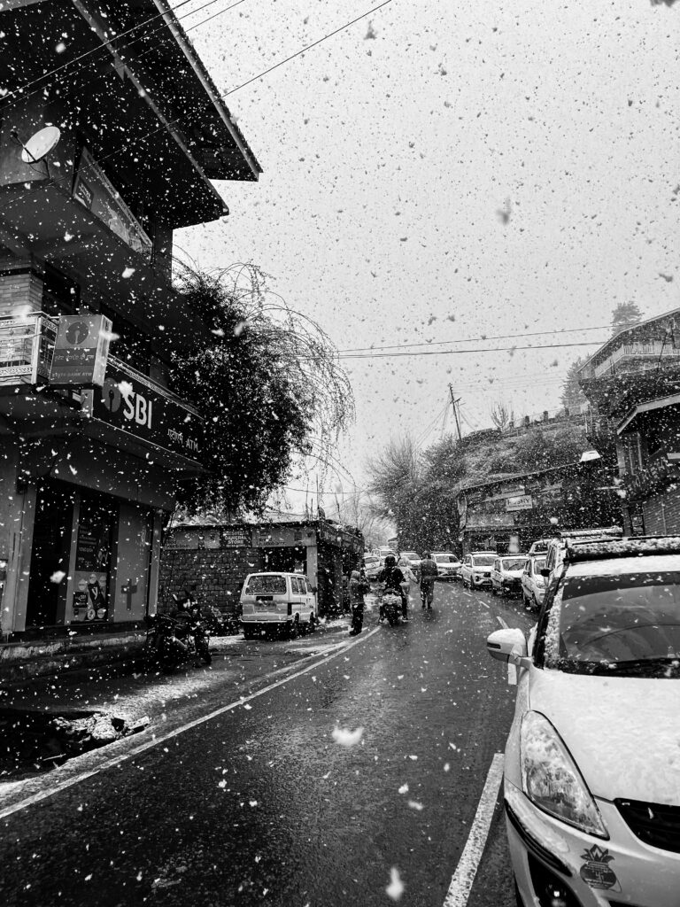Black and white hill-town street in falling snow with parked cars