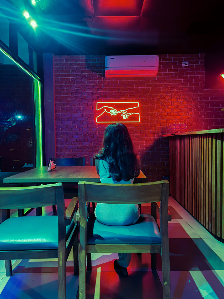 Woman sitting alone at a café table under a red neon sign, lit in teal and red