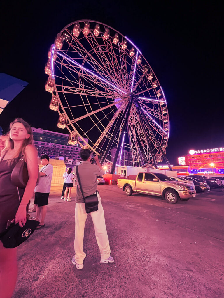 Visitors photographing a lit ferris wheel at a night fair under a purple sky