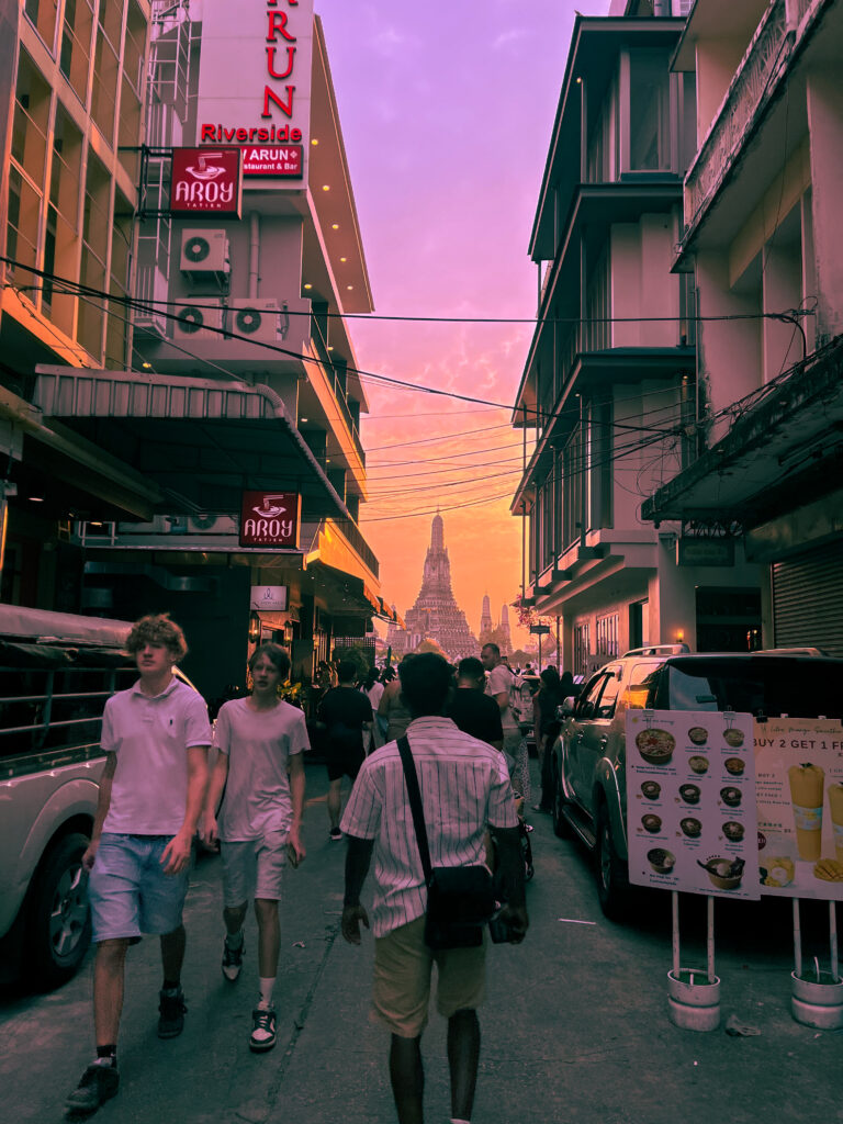 Busy Bangkok street at sunset with the spire of Wat Arun temple at the end of the road