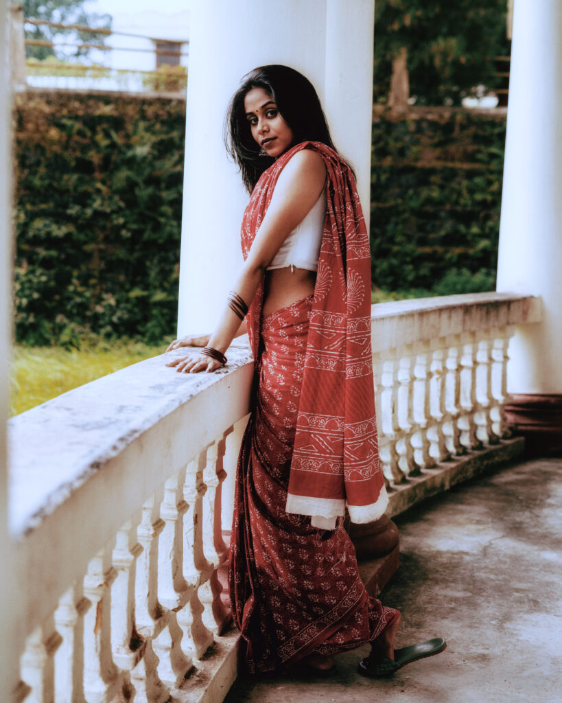 Woman in a red printed saree leaning on a white colonnade balcony