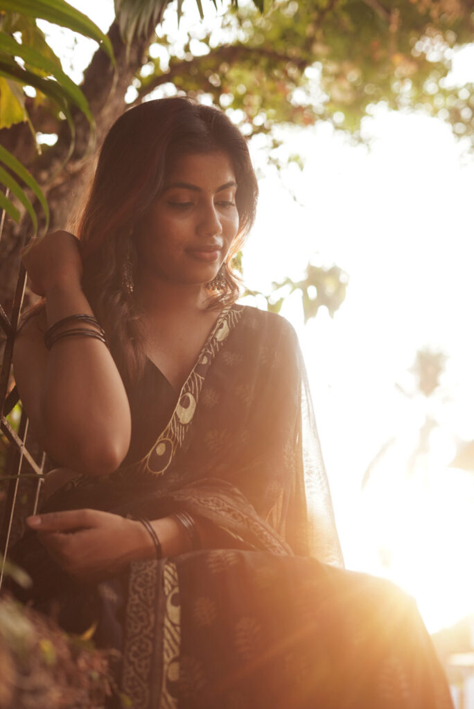 Woman in a black printed saree seated in warm evening light among leaves